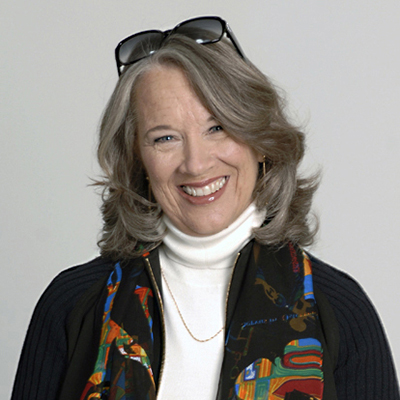 Headshot of Joanne Leedom-Ackerman, a smiling White woman with sunglasses perched on top of her head and wearing a white top and black jacket