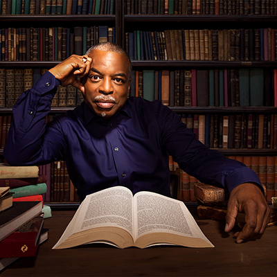 Headshot of LeVar Burton, a Black man in a dark blue collared shirt sitting at a desk in a library with a book open in front of him