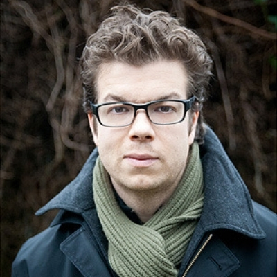 Headshot of author Ben Lerner, a White man wearing glasses, a green scarf, and a blue coat