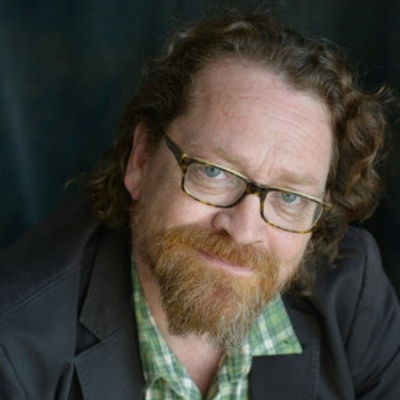 Headshot of author Dan Chaon, a White man with glasses and a beard wearing a green patterned shirt and a black blazer