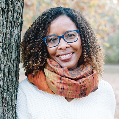 Headshot of Lauren Francis-Sharma, a smiling Black woman wearing glasses, a patterned scarf, and a white top