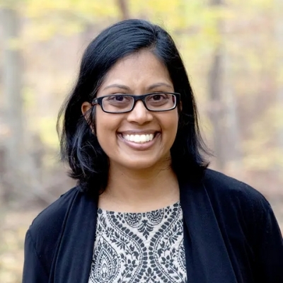 Headshot of author Mathangi Subramanian, a smiling South Asian woman wearing glasses and a black sweater