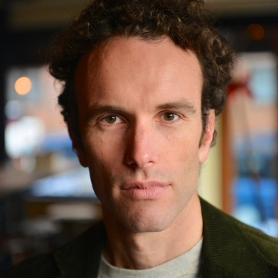 Headshot of White man, Elliot Ackerman, with short curly brown hair wearing a green cardigan and gray top