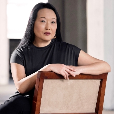 Headshot of Asian woman, Jung Yun, sitting on a chair with long black hair wearing gold dangly earrings and a black dress