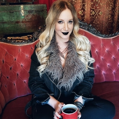 Headshot of author Leigh Bardugo, a person with long blonde hair wearing dark makeup and a black furry jacket, holding a mug of coffee