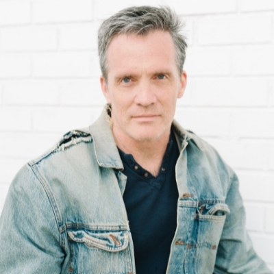 A headshot of Louis Bayard, a White man with short hair wearing a blue top and denim jacket