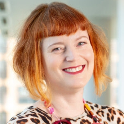 Headshot of Petra Mayer, a person smiling at the camera with short orange hair wearing a colorful necklace and printed top