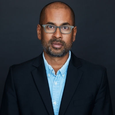 Headshot of Rumaan Alam, a person with close-cropped hair and stubble wearing glasses, a blue collared shirt, and a black suit jacket