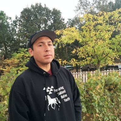Headshot of author Tommy Orange, a person wearing a cap and a black hoodie that says 'Preparing Oakland Native Youth'