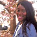 Headshot of Lisa Page Writing Workshops Fellow Faith Angel Campbell, a person with long black hair smiling at the camera wearing a purple jacket and white top against a background of flowers