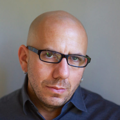 Headshot of Alexi Zentner, a White man wearing glasses, earrings, and a dark collared top