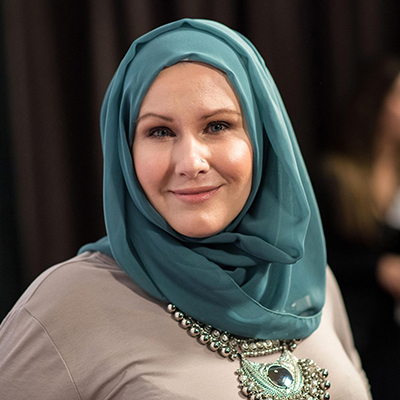 Headshot of author G. Willow Wilson, a smiling woman wearing a blue headscarf and a gray top