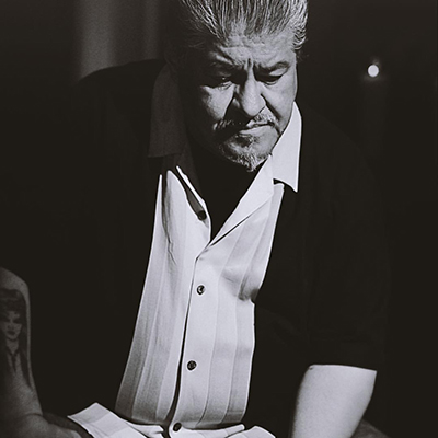 Black and white headshot of author Luis J. Rodriguez, a Xicanx person wearing a white collared shirt and black jacket