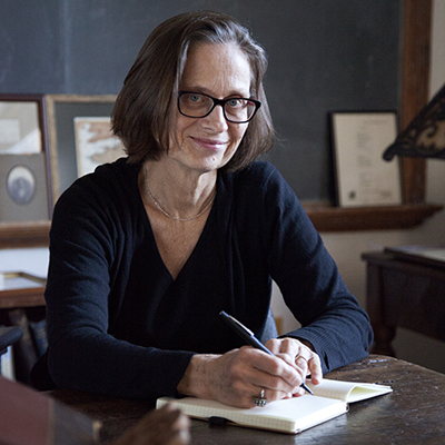 Headshot of writer Lydia Davis, a White woman wearing glasses and sitting at a desk, writing, smiling into the camera