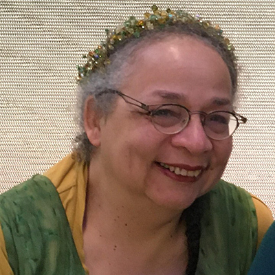 Headshot of author Nisi Shawl, a Black woman wearing glasses and a floral headband, smiling at the camera