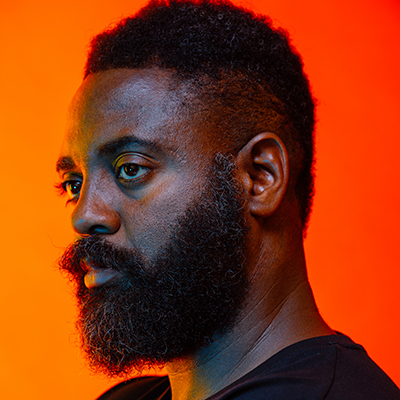 Headshot of author Reginald Dwayne Betts, a Black man with a beard against an orange background