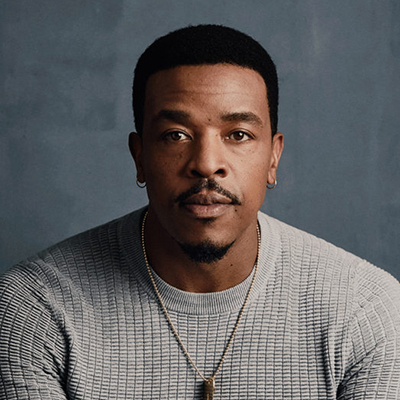 Headshot of actor Russell Hornsby, a Black man wearing earrings, a necklace, and a gray sweater