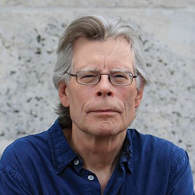 Headshot of author Stephen King, a White man wearing glasses and a blue collared shirt